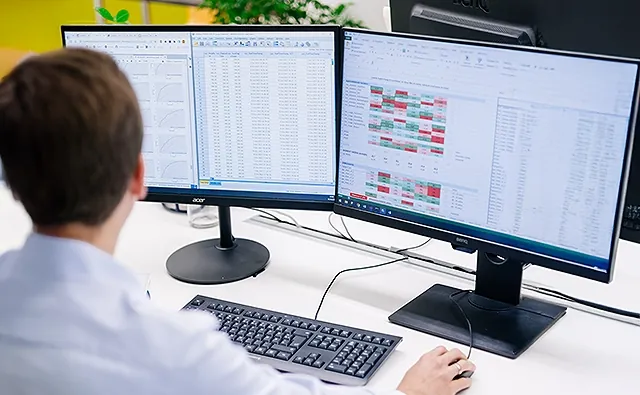 Thomas Schuster arbeitet an zwei großen Computerbildschirmen, auf denen umfangreiche Excel-Tabellen mit Diagrammen und farblich markierten Daten zu sehen sind.