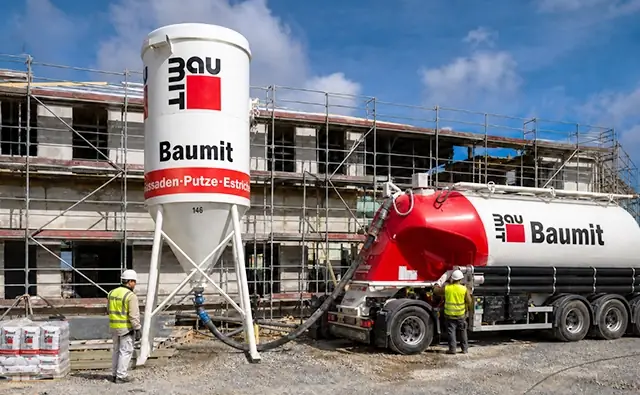 Baumit-Silo und Baumit-LKW auf einer Baustelle vor einem eingerüsteten Gebäude, während Bauarbeiter Material verarbeiten. Bild steht für Baustoffe, Bauprozesse und professionelle Fassaden- und Putzlösungen.