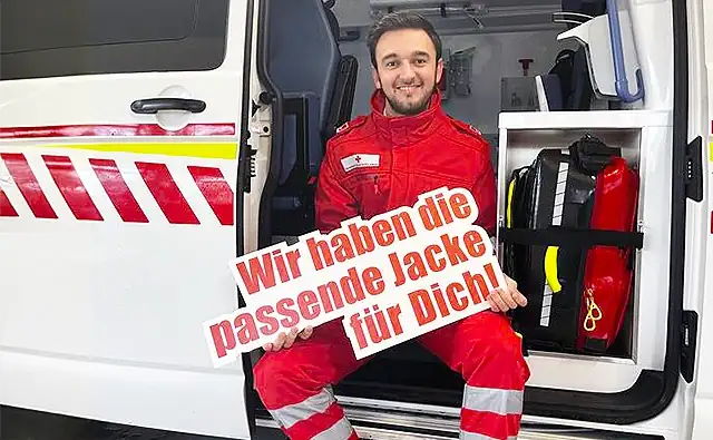 Ein freiwilliger Rettungshelfer in roter Einsatzkleidung sitzt in der geöffneten Seitentür eines Rettungswagens und hält ein Schild mit der Aufschrift 'Wir haben die passende Jacke für Dich!' in den Händen. Im Hintergrund ist Ausrüstungsmaterial im Fahrzeug sichtbar.
