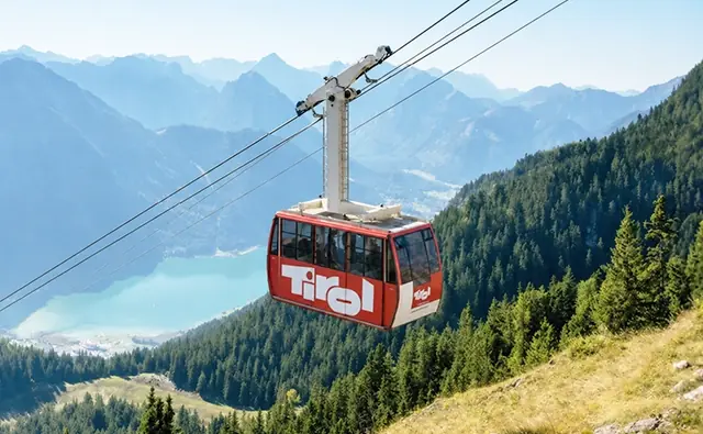 Eine Seilbahn mit Tirol-Branding beim der Bergfahrt - mit Blick auf einen türkisfarbenen Bergsee und bewaldete Alpen.