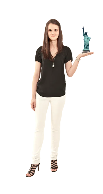 Mag. Birgit Schrenk trägt schwarze Bluse und weiße Hose und balanciert die Freiheitsstatue in Miniaturform auf ihrer Hand – ihre Liebe zu Reisen ist unverkennbar.
