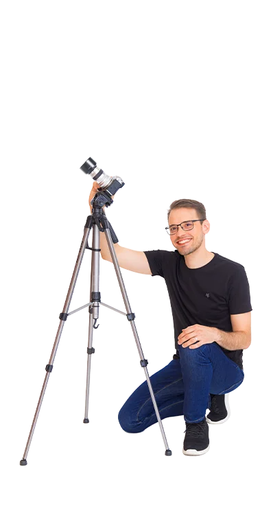 Mag. Daniel Winzer kniet lächelnd neben einem Stativ mit Kamera – mit Leidenschaft für Fotografie und den perfekten Moment.