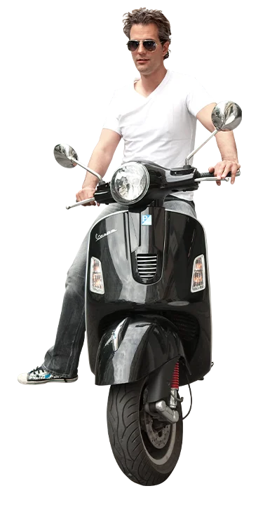 Mag. Erik Stadler trägt ein weißes T-Shirt und Sonnenbrille und fährt lässig auf einer schwarzen Vespa – ganz im Stil von Dolce Vita.