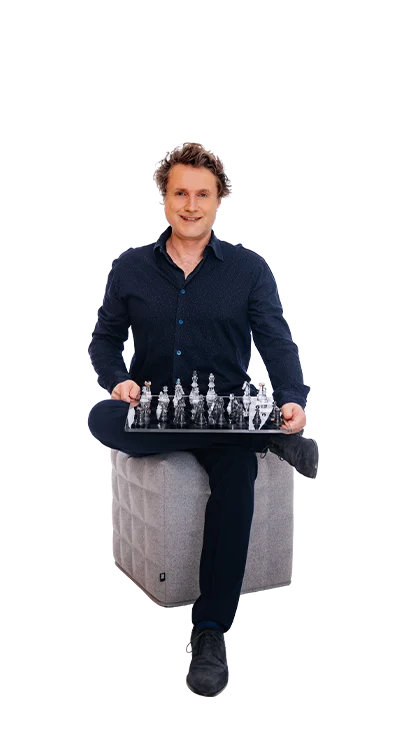 Dipl.-Wi.-Ing. Dr. Gereon Friederes sitzt entspannt auf einem grauen Hocker und hält ein Schachbrett mit transparenten Figuren – strategisch, fokussiert und mit einem Lächeln.
