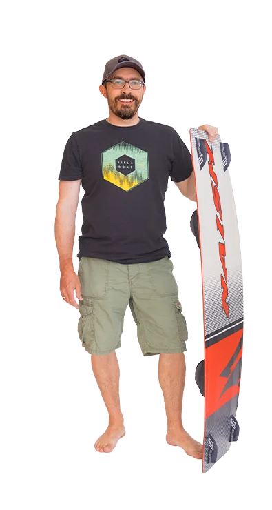 Mag. Gerhard Schüller steht barfuß, trägt ein T-Shirt mit Surfmotiv und hält ein Kiteboard – sportlich, entspannt und bereit für Wind und Wellen.