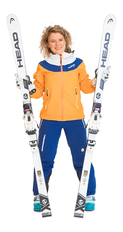 Mag. Heidi Thyri steht in kompletter Skimontur mit orange-blauer Jacke und hält zwei weiße Skier fest in den Händen – bereit für die Piste und jede Menge Schneeabenteuer.