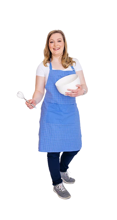 Katharina Beck, MSc (WU) trägt eine blaue Küchenschürze und hält lächelnd eine weiße Rührschüssel sowie einen Schneebesen – backfreudig, herzlich und voller Tatendrang.