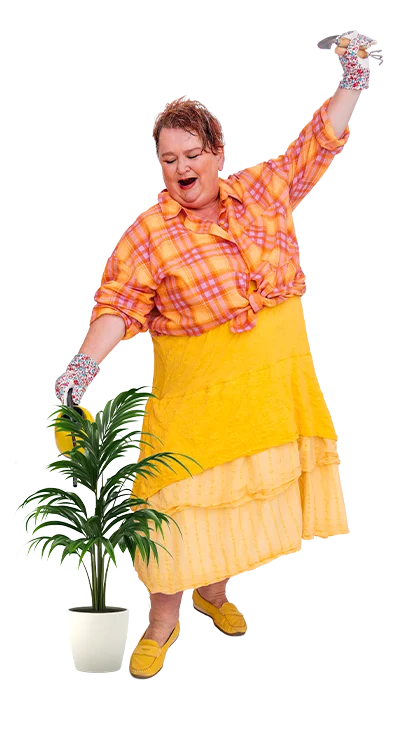 Lydia Croce tanzt fröhlich mit Gummihandschuhen, Gießkanne und bunter Kleidung um eine Topfpflanze herum – lebenslustig, farbenfroh und voller Humor.