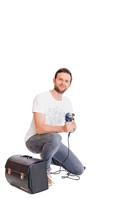 Lucas Pum, MA hockt mit Bohrmaschine in der Hand neben einer Tasche, trägt Jeans und T-Shirt – technikbegeistert, kreativ und fokussiert.