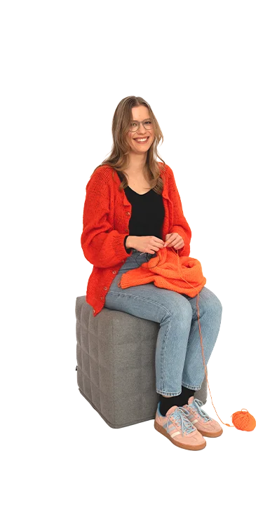 Polina Yaroshchuk, BA MA sitzt mit oranger Strickjacke und Wolle auf einem Hocker, das Strickzeug in der Hand – kreativ, konzentriert und handwerklich geschickt.