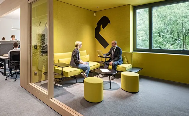 Beratungsgespräch zwischen Bankbetreuer und Kundin in einem modernen, gelb gestalteten Besprechungsraum mit großen Fenstern und Blick ins Grüne. Im Hintergrund ist ein offener Büroraum mit mehreren arbeitenden Personen sichtbar.