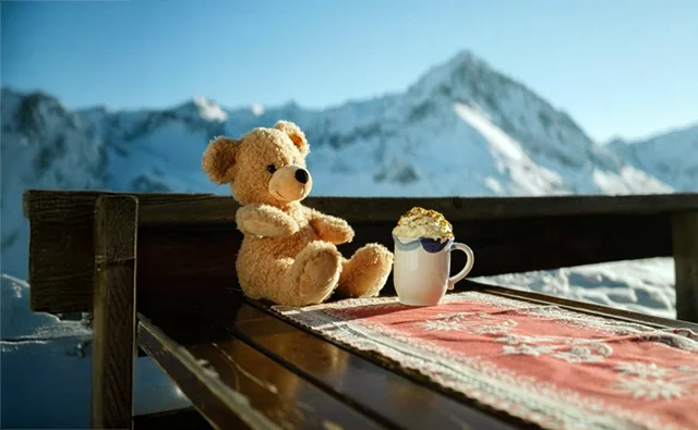 Szene aus der zielgruppenspezifischen Werbekampagne 'Immer für dich da'. Steiff-Teddybär Pauli mit heißer Schokolade auf einer Almhütte vor imposanter Alpenkulisse.