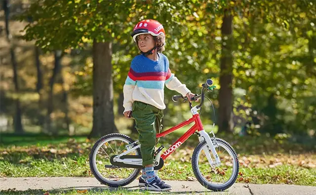 Kind mit rotem Fahrradhelm steht mit einem roten Woom-Kinderfahrrad im Park.