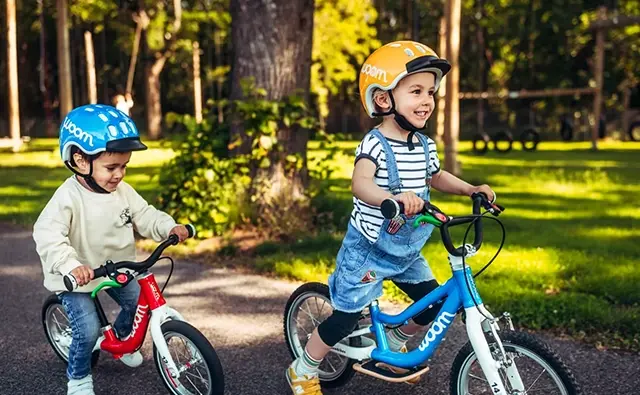 Zwei Kinder fahren fröhlich mit farbenfrohen Woom-Laufrädern durch einen sonnigen Park. Symbolbild für kindgerechte Mobilität, sicheres Fahrvergnügen und durchdachtes Produktdesign der Marke Woom.