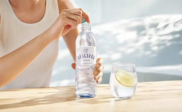 Person öffnet eine Flasche Vöslauer Mineralwasser mit Kohlensäure neben einem Wasserglas mit Zitronenscheibe.