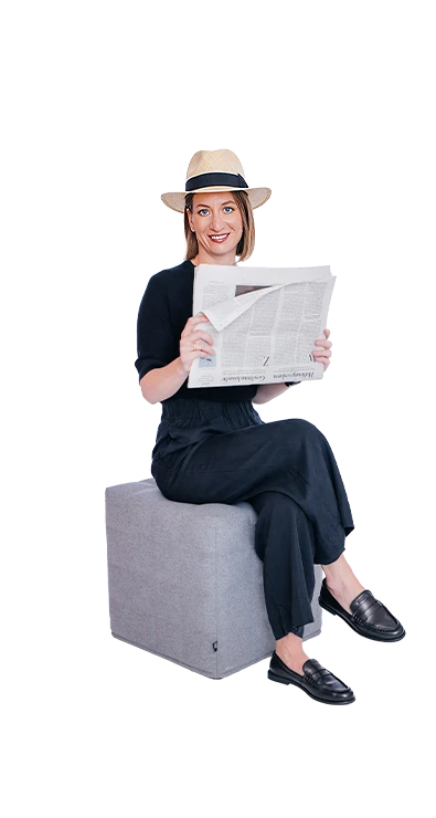 Mag. Barbara Khayat-Platzl, BSc (WU) sitzt mit übergeschlagenen Beinen und Zeitung in der Hand lächelnd auf einem Hocker - entspannt, stilvoll und immer gut informiert.