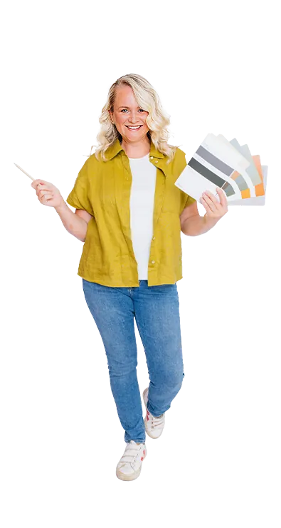 Mag. Kerstin Hofer steht mit Farbfächer und Pinsel in der Hand und lächelt begeistert in die Kamera - kreativ, lebensfroh und voller Gestaltungslust.