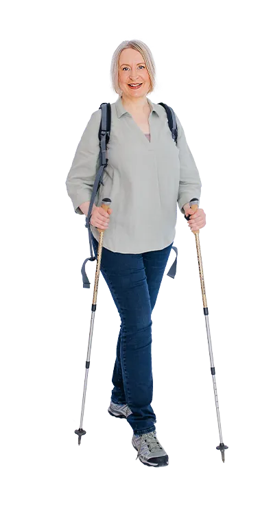 Mag. Sonja Ehrenberger geht mit Wanderstöcken und Rucksack lächelnd auf die Kamera zu - naturverbunden, aktiv und voller positiver Energie.